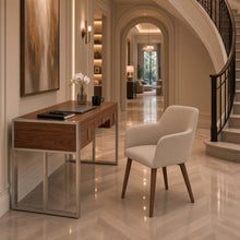 Load image into Gallery viewer, A modern home office setup featuring the Cira wooden desk with a chrome metal frame and a cream upholstered armchair, set in a luxurious hallway with a staircase and polished herringbone tile floors.