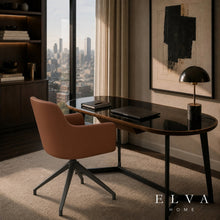 Load image into Gallery viewer, A modern home office scene featuring an Elva swivel chair in leather finish, paired with an oval desk that has a black glass top and dark metal legs. The setup is styled with a dome lamp, laptop, and books, set against a warm beige wall with abstract art and a city skyline visible through a large window.