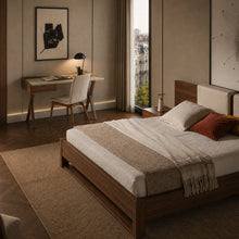 Load image into Gallery viewer, A modern bedroom featuring a low wooden platform bed with cream bedding, a matching wooden headboard, and a textured beige throw blanket. The room has warm beige walls, a built-in wooden desk with a white chair, and a large window looking out onto a city view.