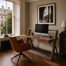 Load image into Gallery viewer, A modern wooden writing desk with a built-in shelf and angled legs, styled in a home office with a brown leather swivel chair, an iMac, and framed wall art.
